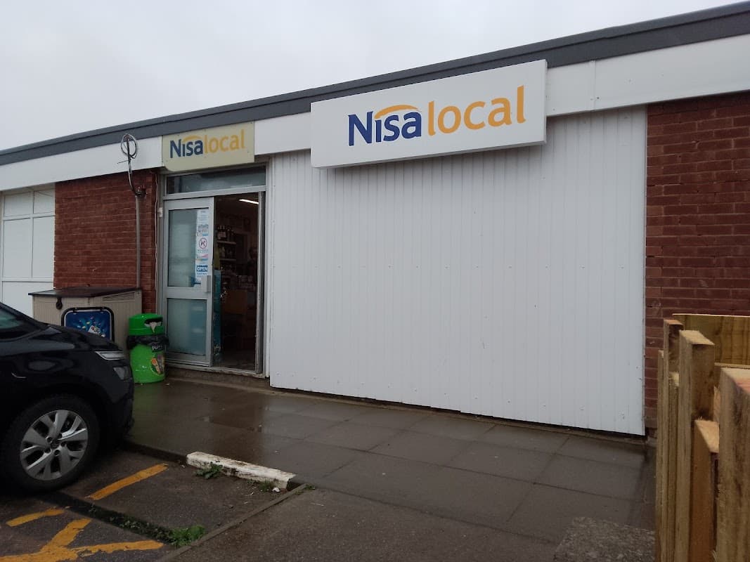 Nisa Local grocery store entrance with a sign, brick wall, and a parked car nearby.