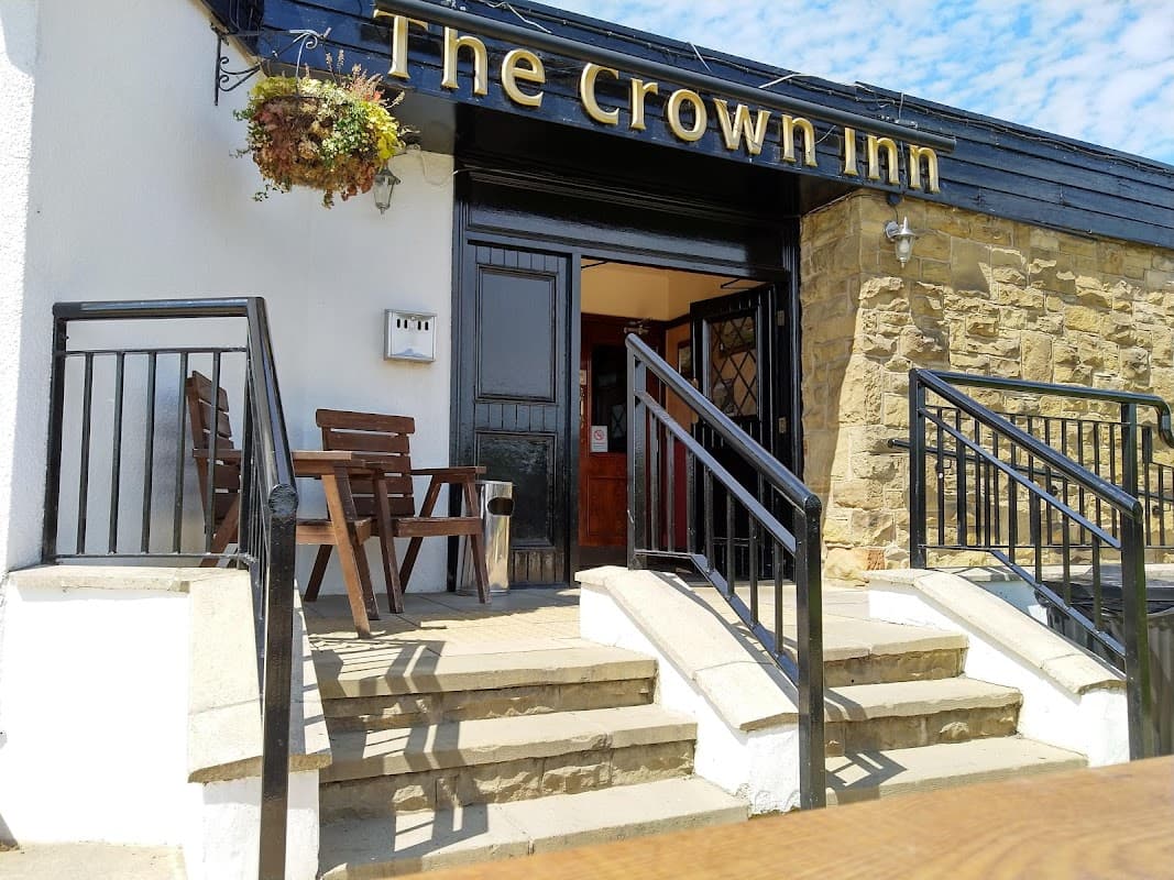 The Crown Inn entrance with wooden chairs, stone steps, and a hanging flower basket under a blue sky.