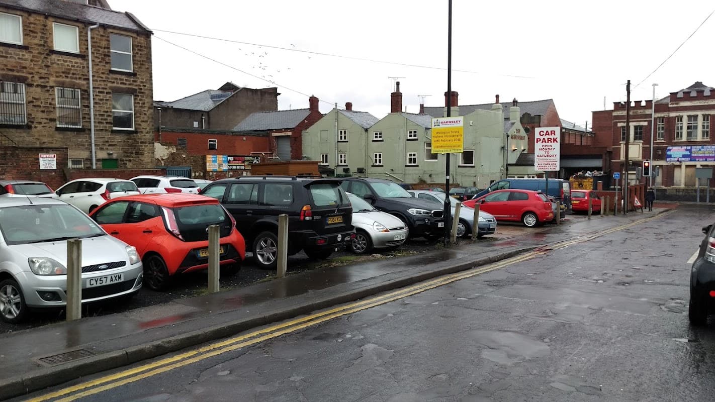 Car park with various parked cars, buildings in the background, and wet pavement under overcast skies.