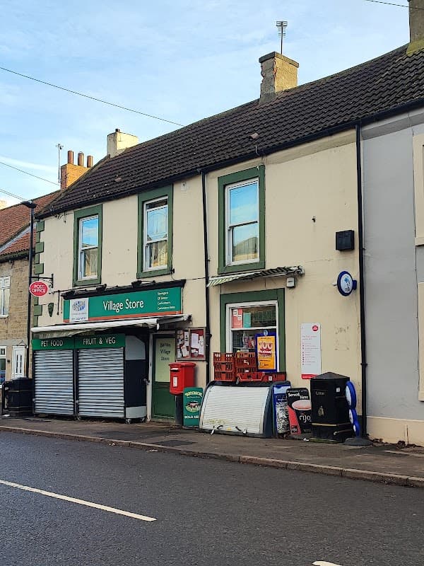 Barton Sub Post Office with green trim, signage for Village Store, and a red post box outside on a quiet street.
