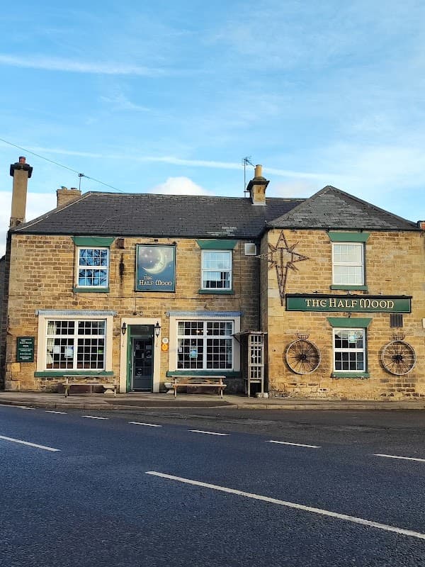 Traditional stone pub with green window frames, wooden benches, and a sign reading "The Half Moon" in Barton, Yorkshire.