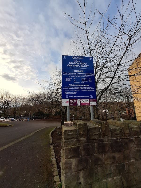 Sign for Station Road Car Park in Batley, detailing parking charges and regulations, surrounded by trees and a stone wall.