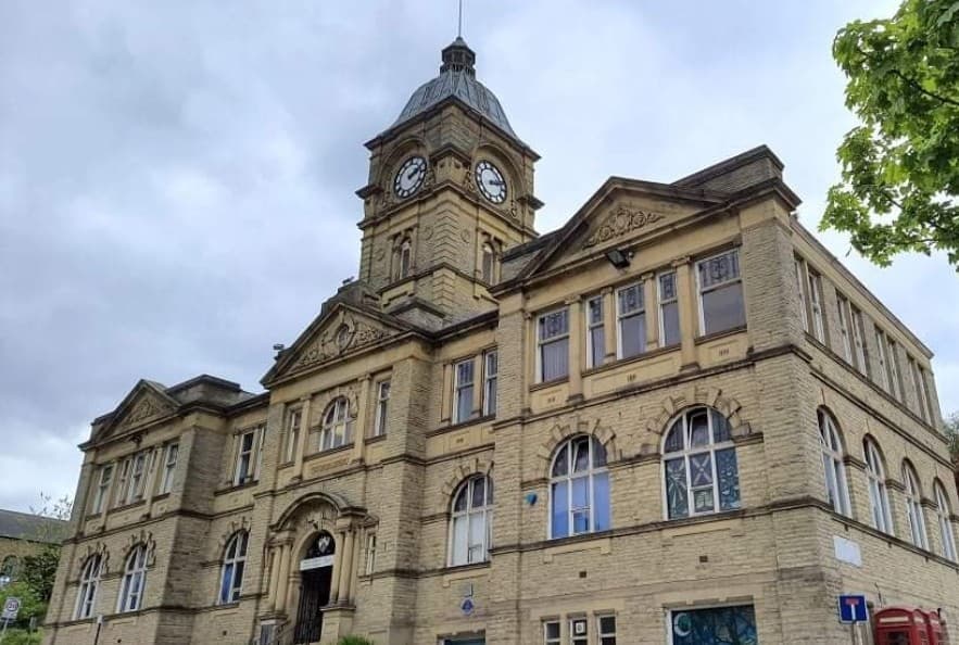 Historic stone building with a clock tower and large arched windows, surrounded by greenery and a cloudy sky.