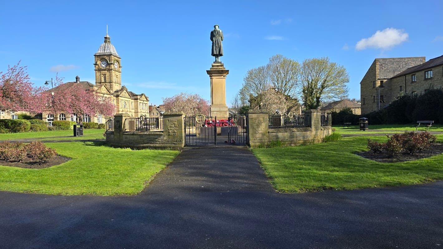 Statue on a pedestal surrounded by greenery and blooming trees, with historic buildings in the background.