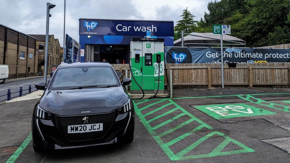 Black Peugeot 208 parked in front of a Be.EV charging station and a car wash facility in Batley, Yorkshire.