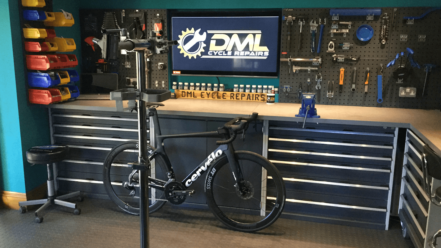 DML Cycle Repairs shop with a bike on a stand, tools, and a display screen against a teal wall.