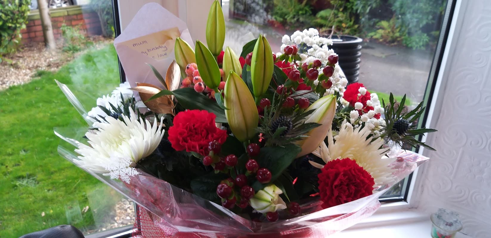 Colorful floral bouquet featuring red and white flowers, green leaves, and decorative elements in a window setting.