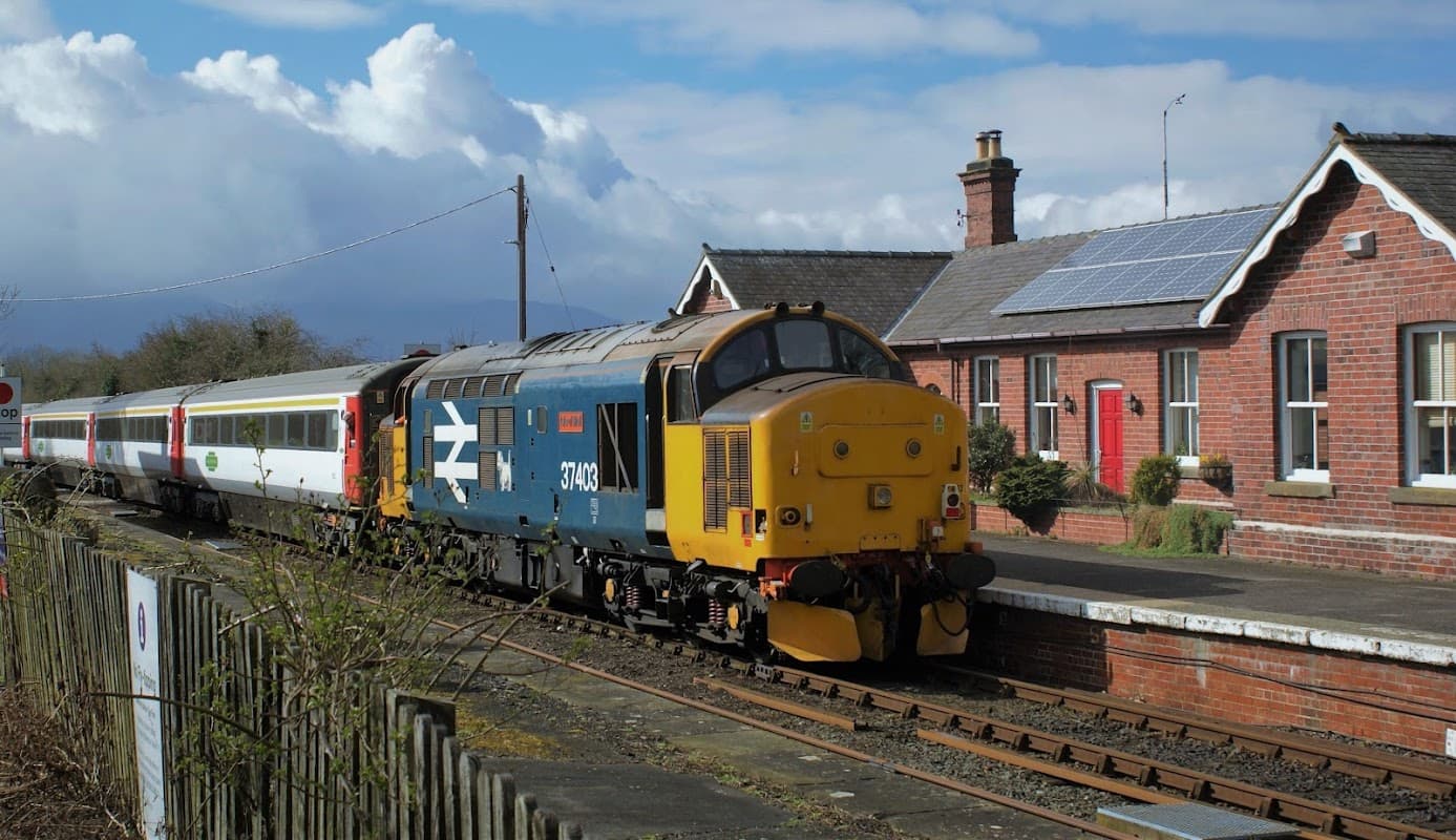 Train at Battersby station with a red-brick building and solar panels under a partly cloudy sky.