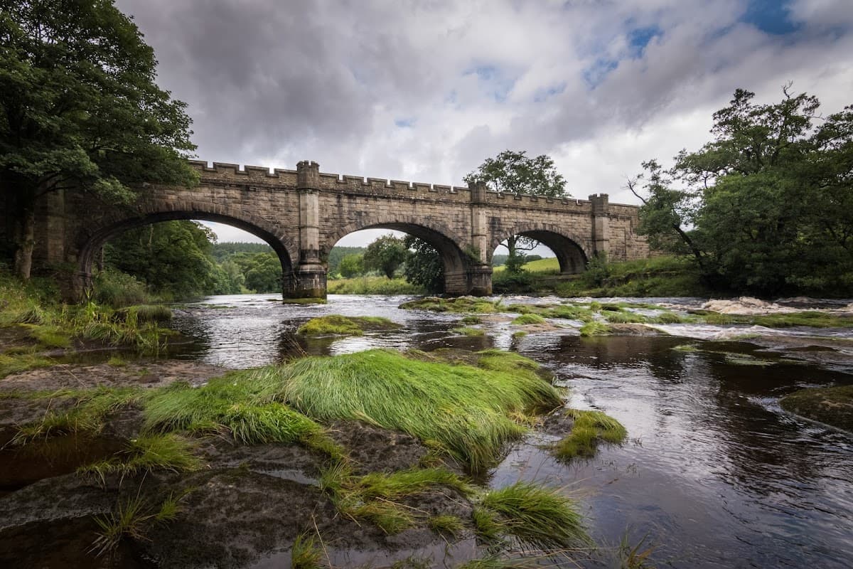 Stone bridge arching over a river, surrounded by lush greenery and cloudy skies in Beamsley, Yorkshire.