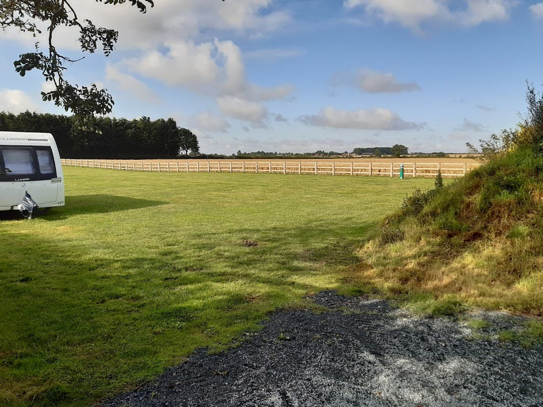 Expansive green field at McKies Caravan Park, with a caravan and wooden fence under a partly cloudy sky.