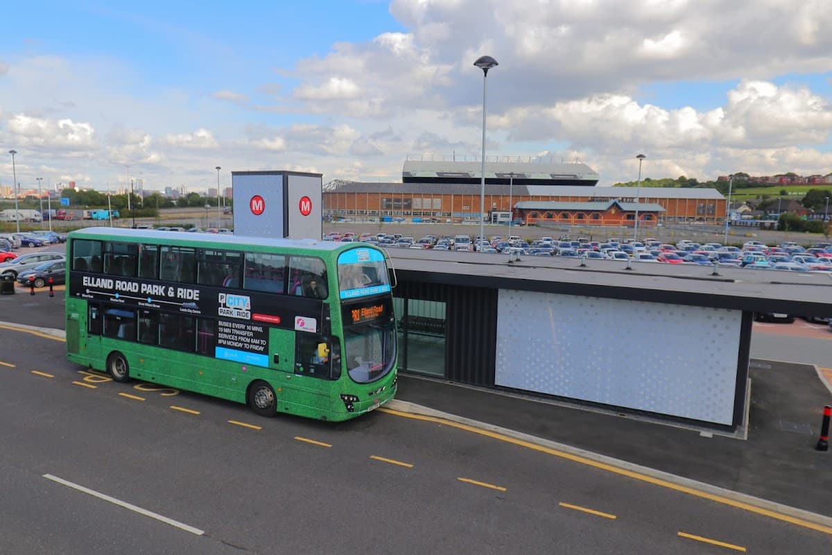 Green bus at Elland Road Park & Ride, with a large parking lot and a stadium in the background under a cloudy sky.