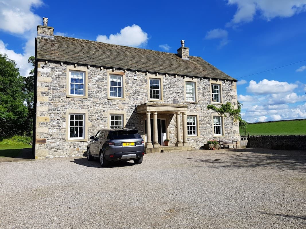 Stone building with a grand entrance, surrounded by greenery and a gravel driveway, featuring a parked car in front.
