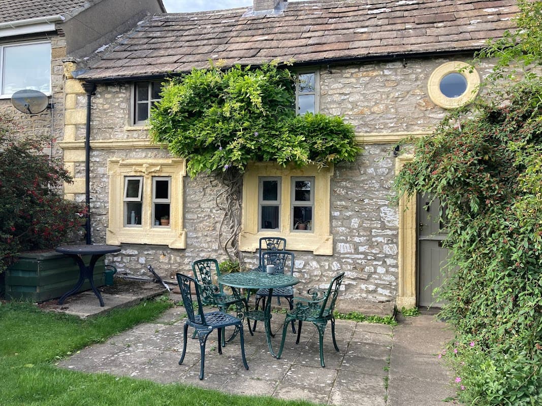 Charming stone cottage with yellow accents, a vine-covered entrance, and a small patio set in a grassy area.
