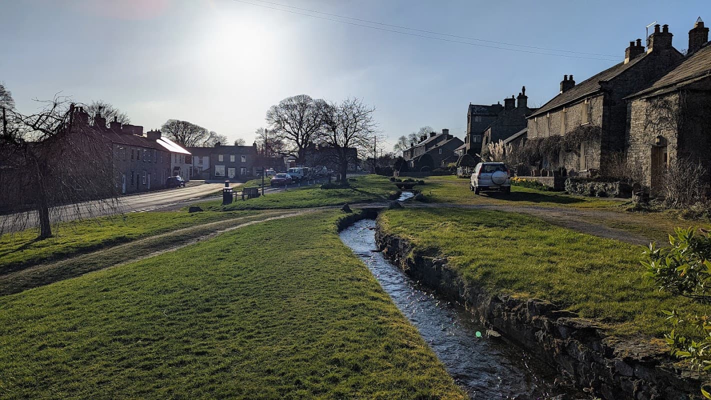Cherry Tree Cottage alongside a grassy area and stream, with stone houses and trees under a bright sky in Bellerby.