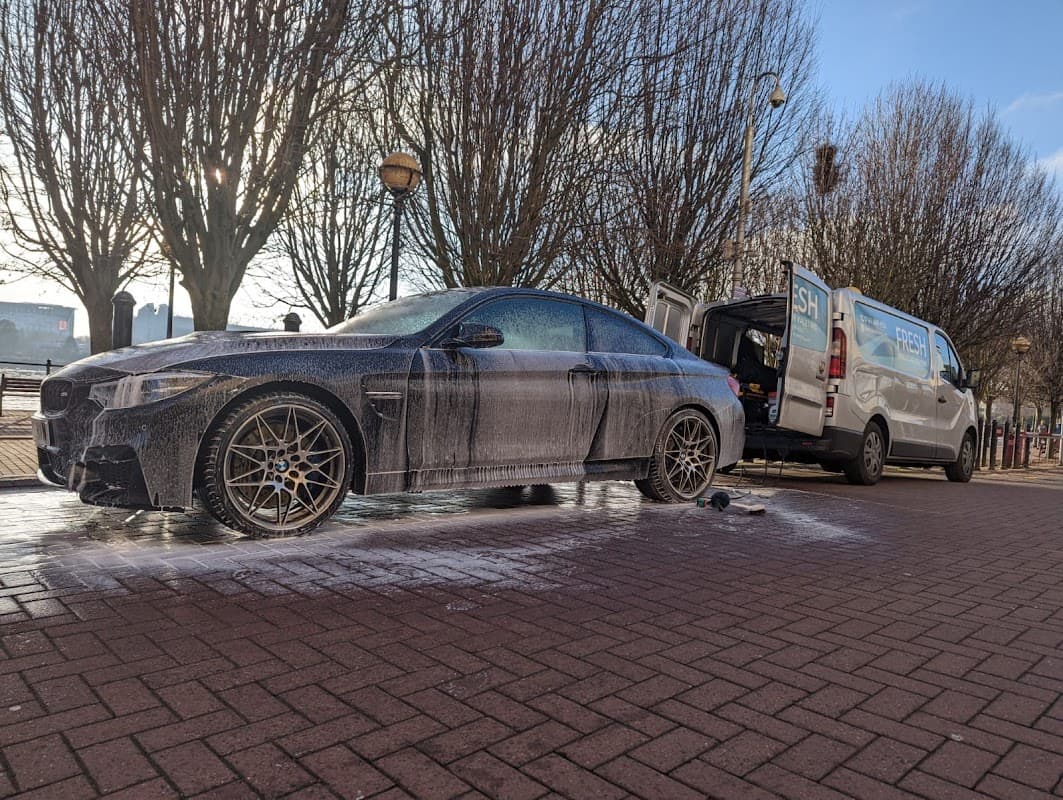 A black car covered in soap sits on a brick street, with a van open nearby and trees in the background.