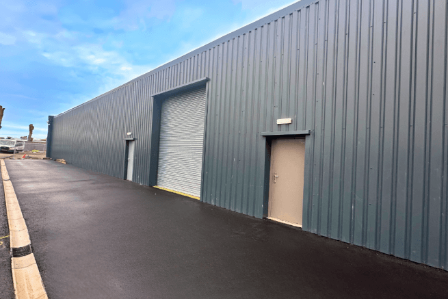 Gray metal building with a large roller door and two smaller doors, set on a freshly paved driveway.