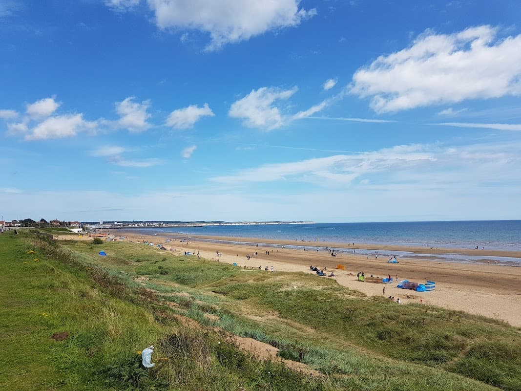 Wide sandy beach with people enjoying the sun, green grass in the foreground, and a clear blue sky with clouds.