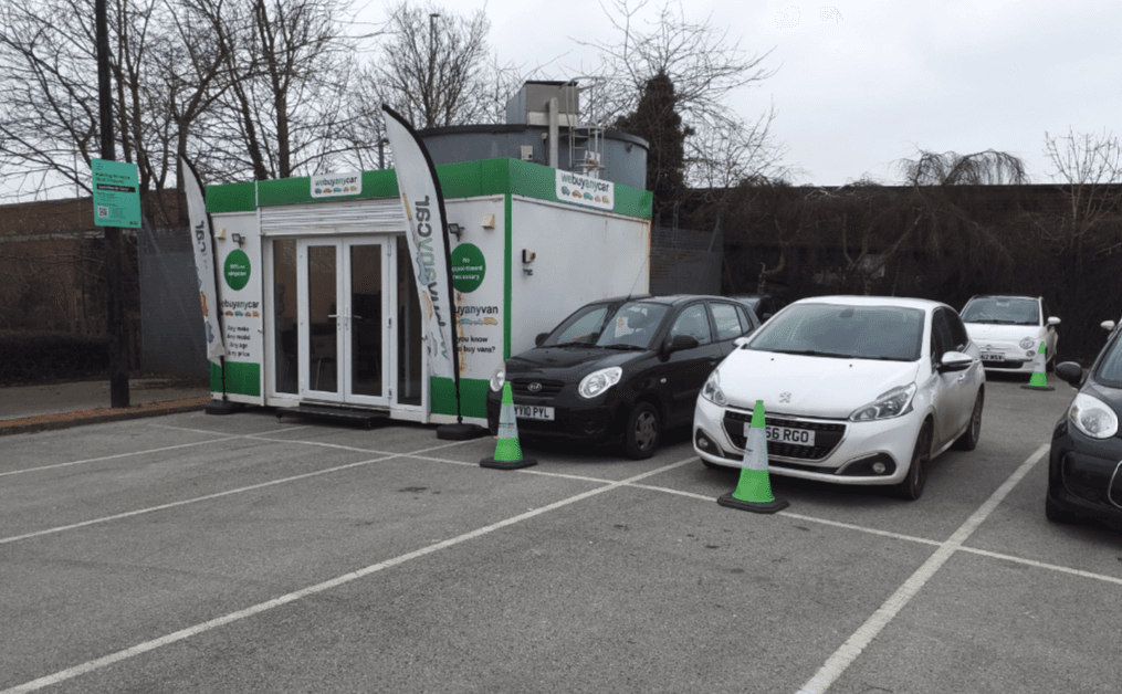 "We Buy Any Car" office with flags, parked cars, and green cones in a lot in Bessingby, Yorkshire.