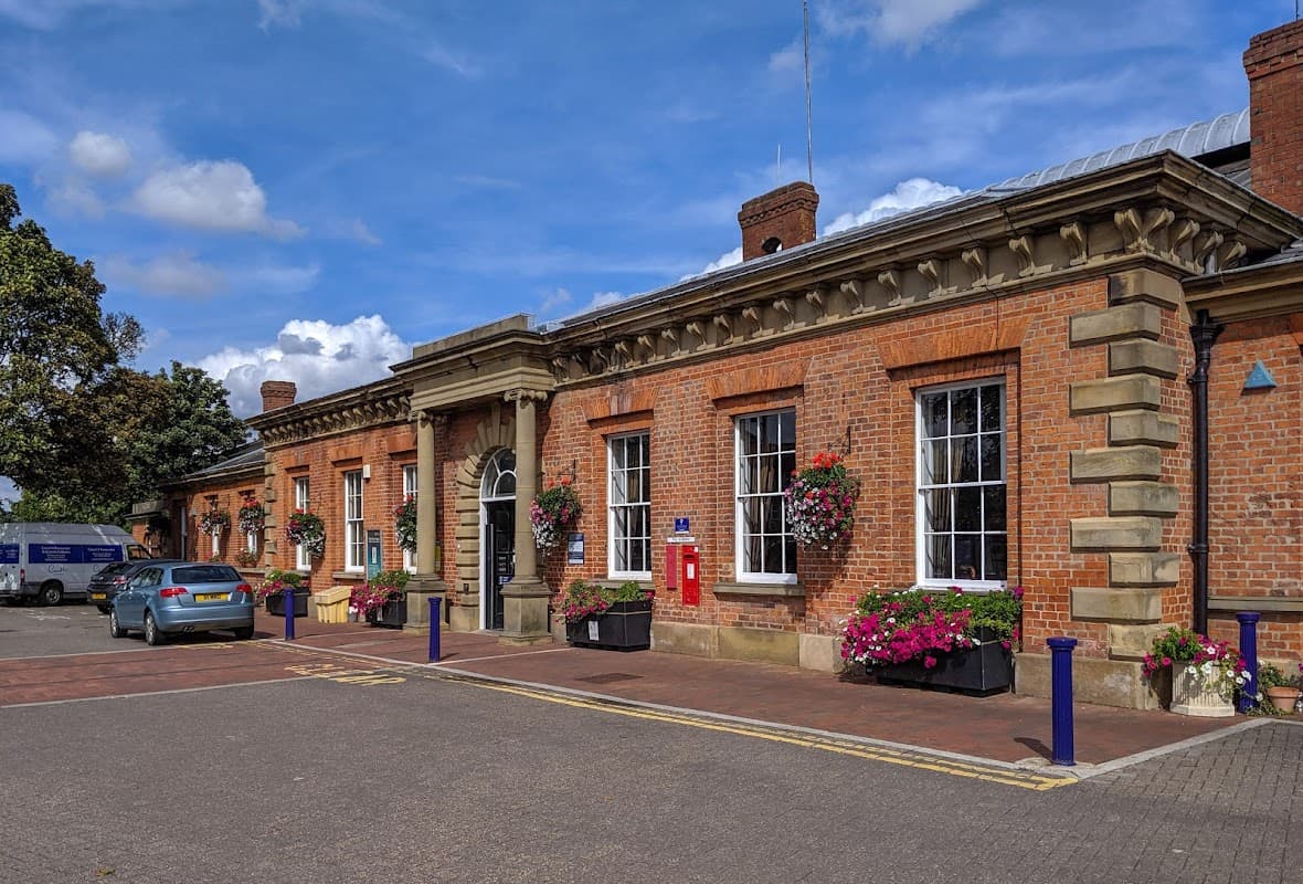 Brick building with large windows, flower planters, a red post box, and parked cars under a blue sky.