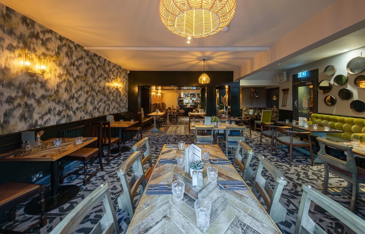 Stylish bar interior with wooden tables, green seating, patterned carpet, and decorative lighting.