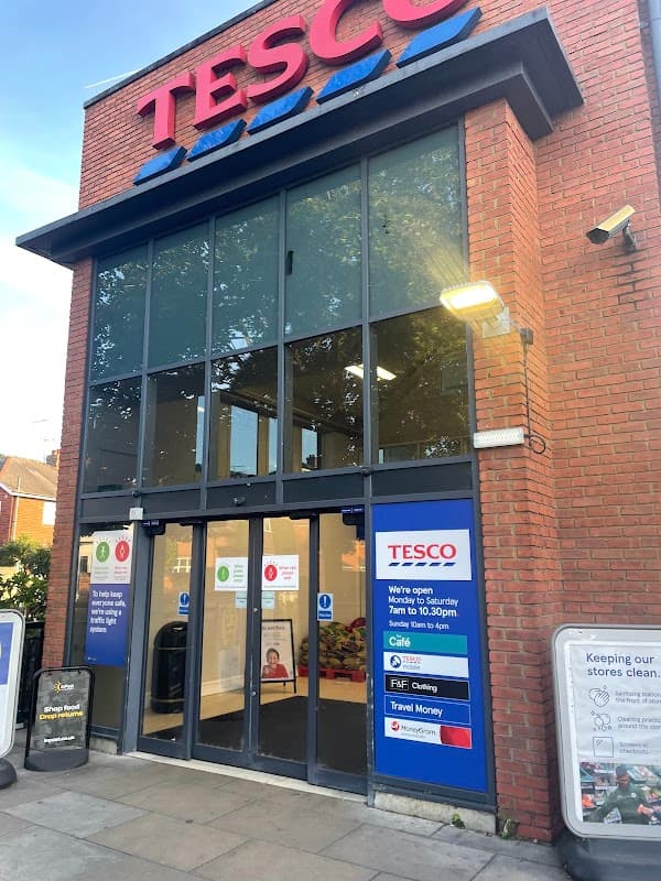 Tesco Superstore entrance with large glass doors, red brick facade, and signage for opening hours and services.