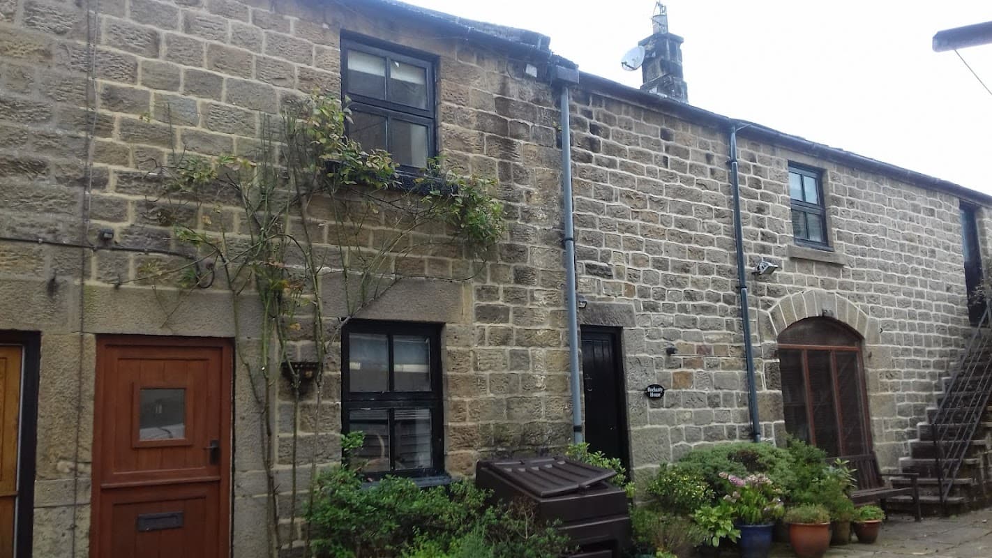 Stone cottage with dark windows, wooden door, and potted plants, set in a rural Yorkshire landscape.