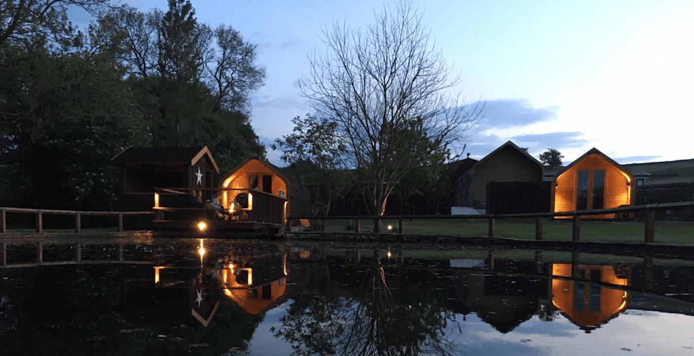 Cozy wooden cabins by a tranquil pond, reflecting trees and evening sky in Bewerley, Yorkshire.