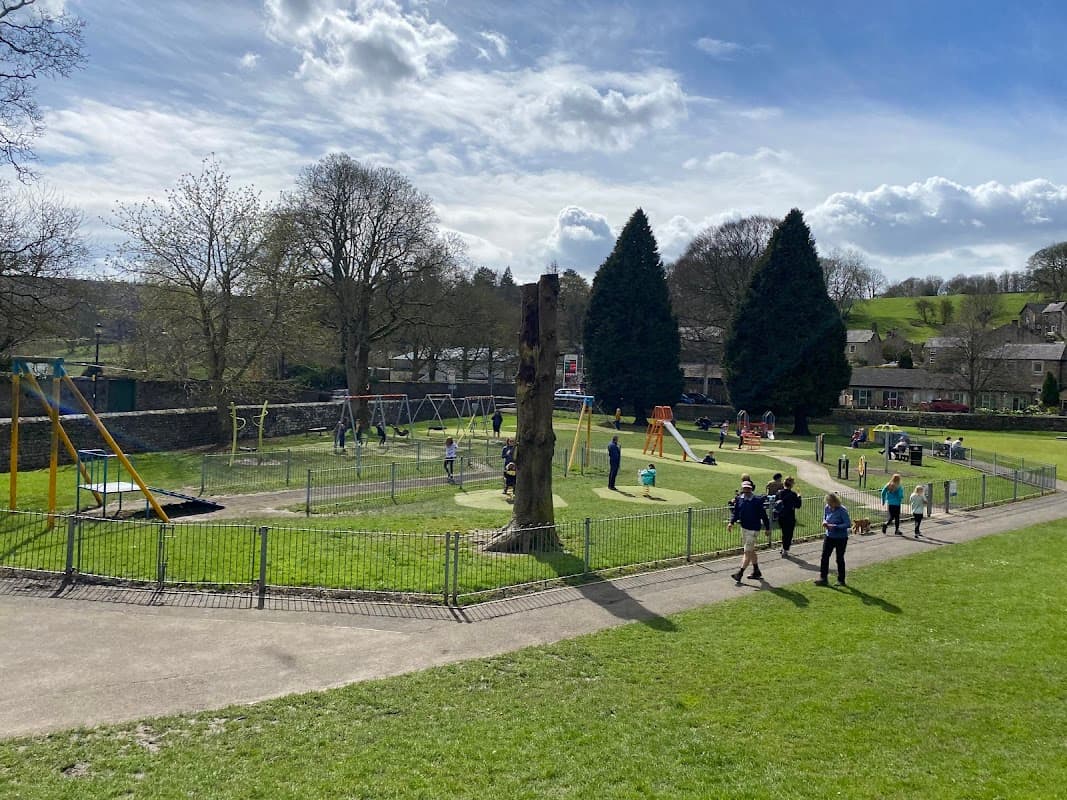 Playground with swings, climbing structures, and children playing on grass, surrounded by trees and a stone wall.