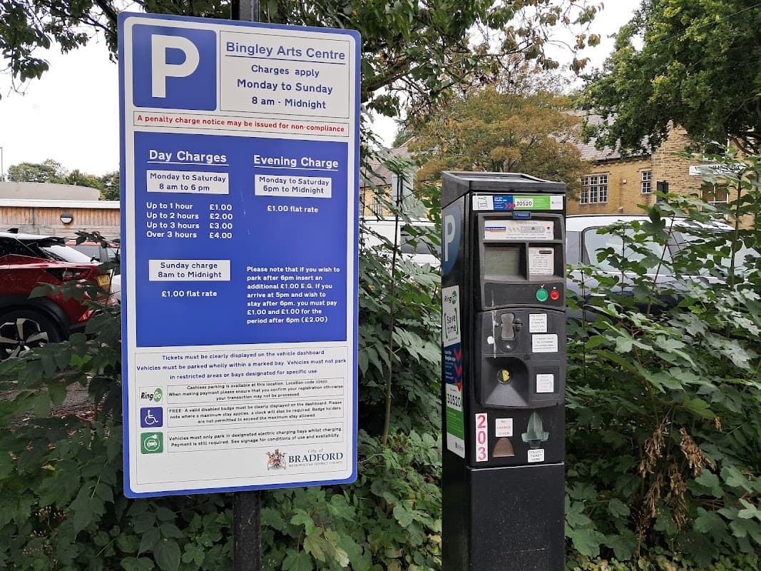 Bingley Arts Centre car park sign with pay & display machine, surrounded by greenery and parked cars.