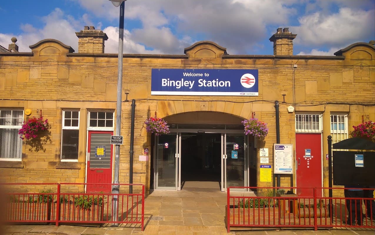 Welcome sign at Bingley Station, with red doors, flower baskets, and a clear blue sky.