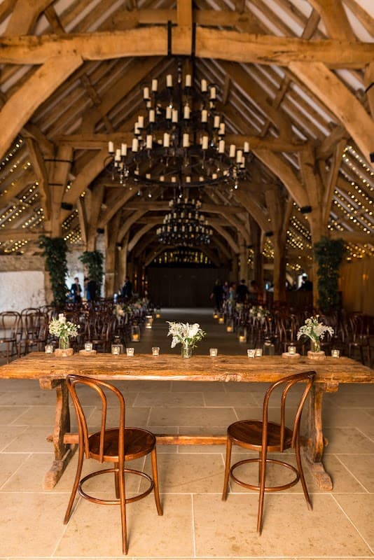 Rustic venue with wooden beams, chandeliers, and floral centerpieces on a long table, surrounded by chairs.