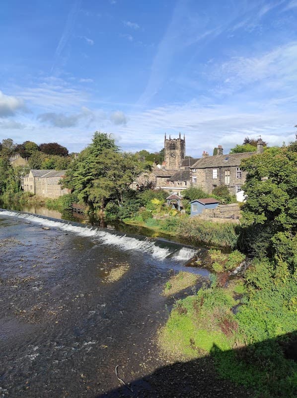 Main Street Car Park in Bingley with a view of a river, trees, and historic buildings under a blue sky.