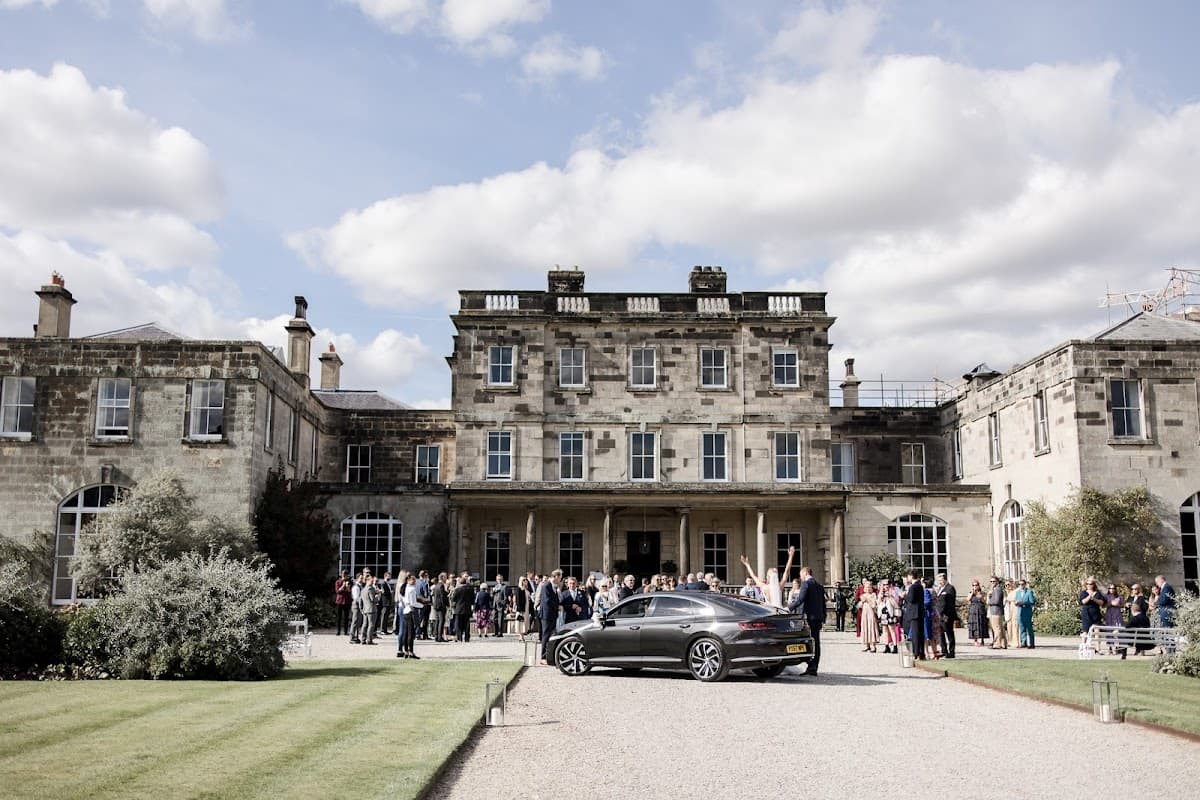 Elegant mansion with guests gathered outside, lush lawn, and a sleek car parked in front under a cloudy sky.