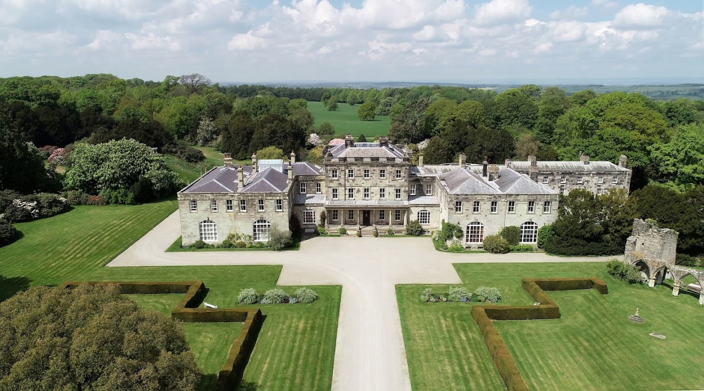 Elegant stone mansion surrounded by manicured lawns and lush greenery under a bright blue sky.