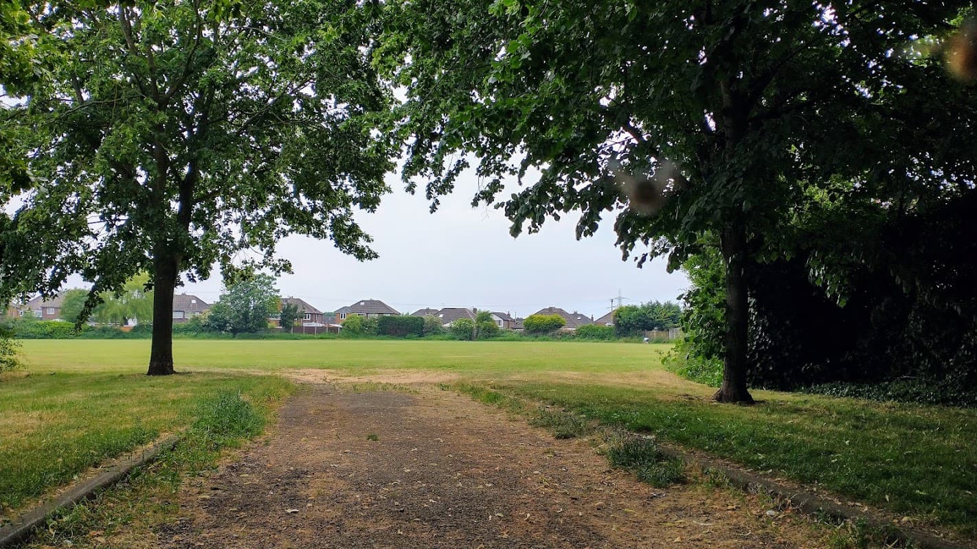 A grassy field framed by trees, with houses visible in the background under an overcast sky.