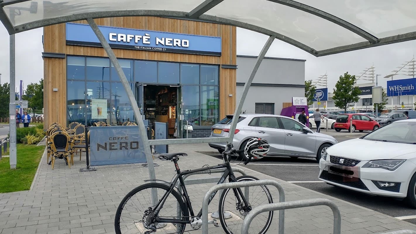 Caffè Nero storefront at Birstall Shopping Park, with a bicycle parked in front and cars in the parking lot.