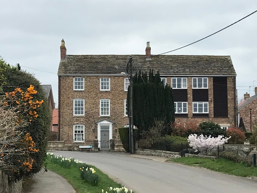 Brick building with multiple windows, surrounded by greenery and flowers, located on a quiet road in Bishop Monkton.