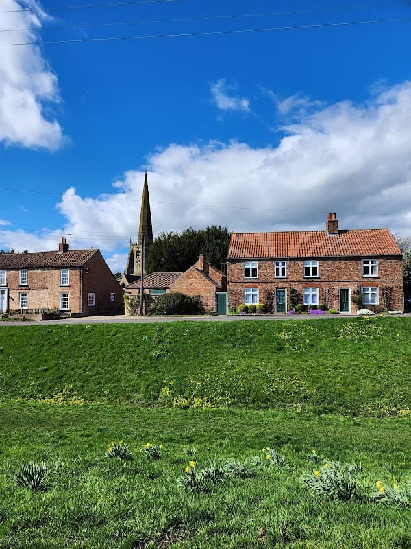 Bishop Wilton Village Hall with stone buildings, a church spire, and a grassy foreground under a blue sky.