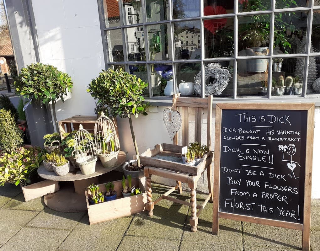 Charming florist display with potted plants, wooden crates, and a humorous chalkboard sign about Valentine's flowers.