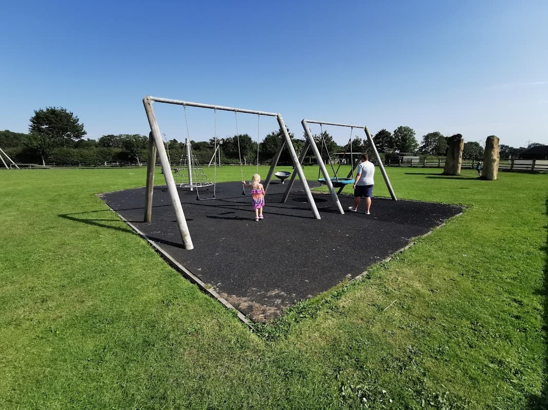A sunny playground with swings, a child playing, and a grassy area surrounded by trees in Borrowby, Yorkshire.