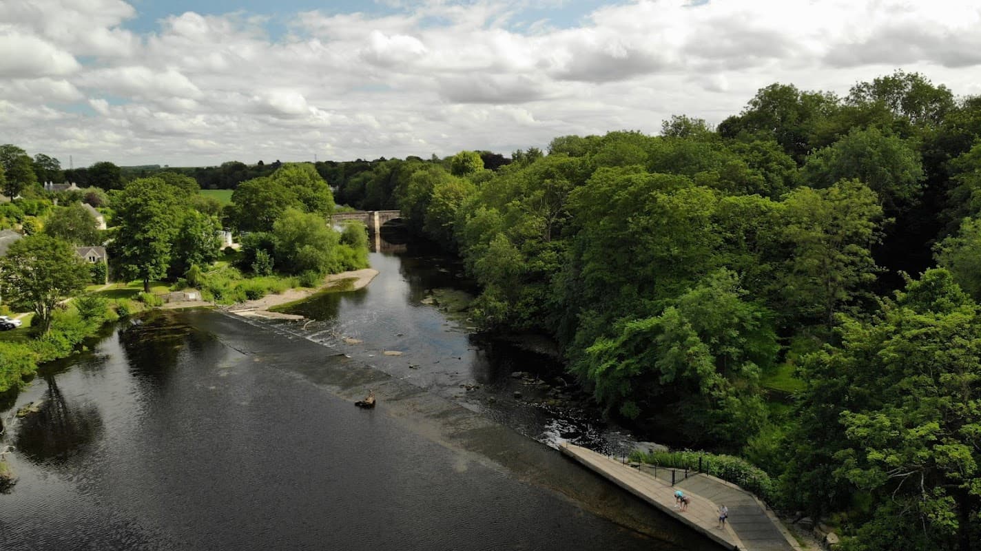 A serene river scene with lush greenery, a pathway, and fluffy clouds overhead in Boston Spa, Yorkshire.