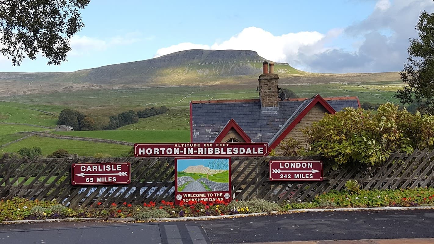Sign for Horton-in-Ribblesdale with distance markers, a quaint building, and a backdrop of rolling green hills.