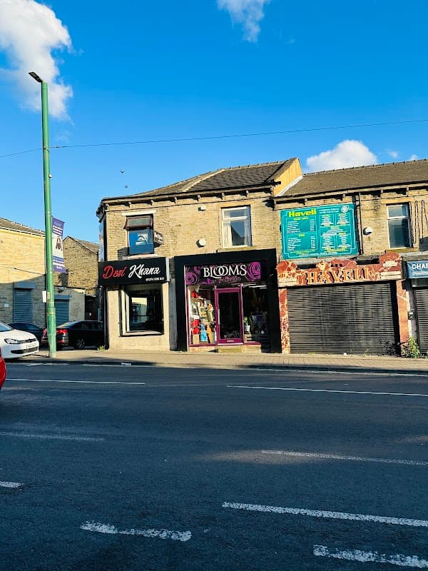 Bright storefront of "Blooms of Bradford" with large windows, colorful signage, and a clear blue sky above.