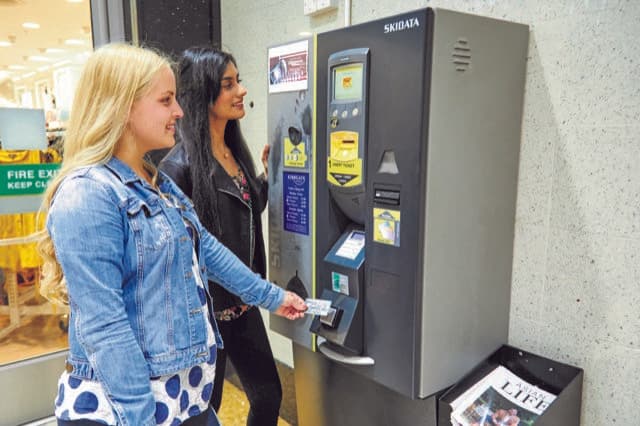 Two young women using a parking payment machine in a car park, with a brochure stand nearby.