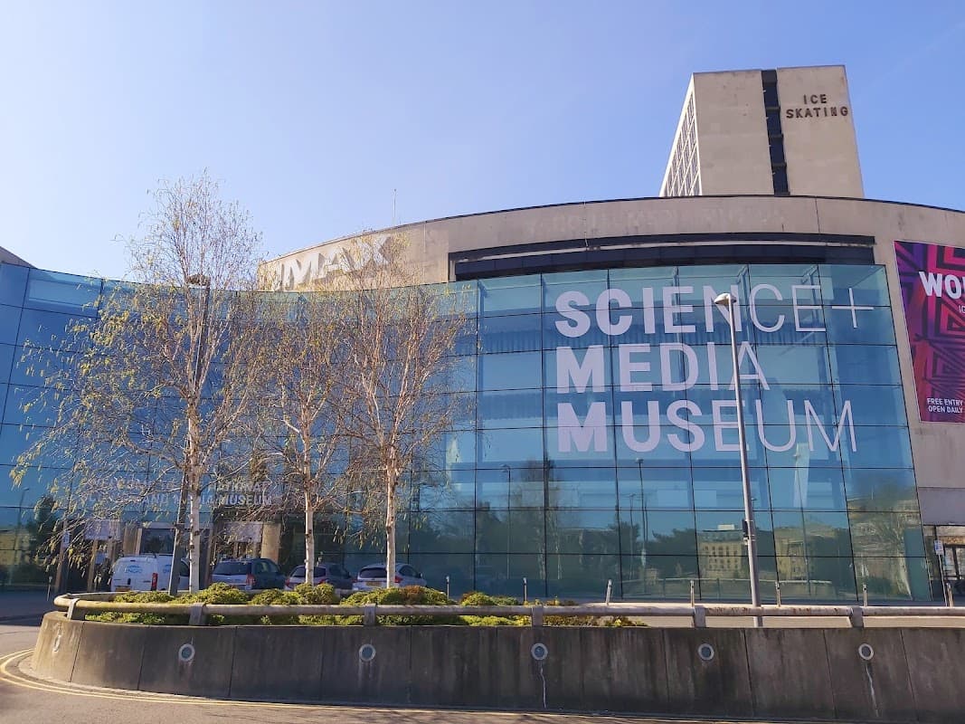Modern glass building with large signage reading "Science + Media Museum," surrounded by trees and urban architecture.