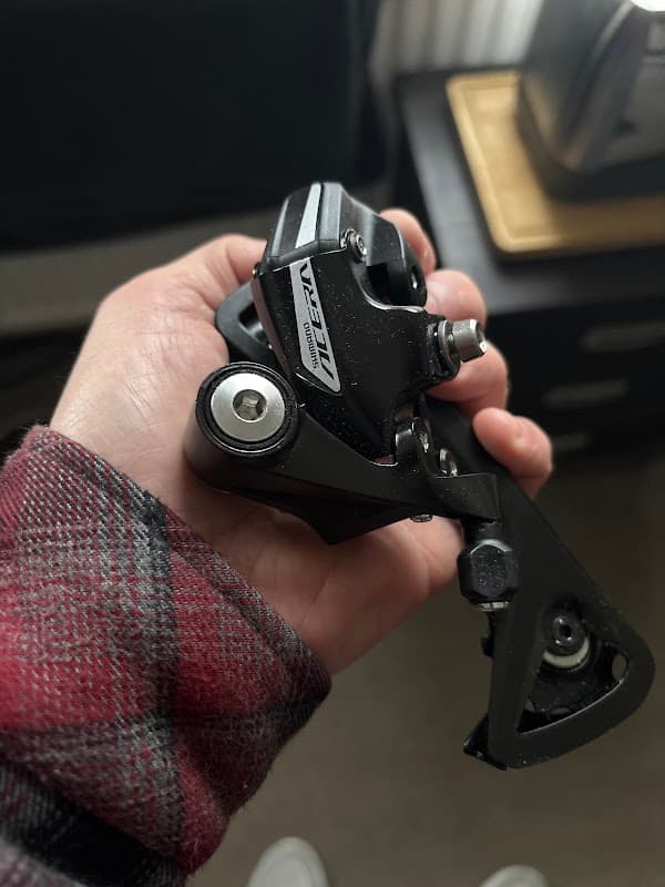 A person holding a black bicycle derailleur with a silver logo, set against a blurred background.