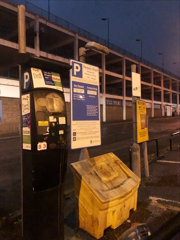 Parking meter and information sign for Rawson Road Car Park, with a large parking structure in the background at dusk.