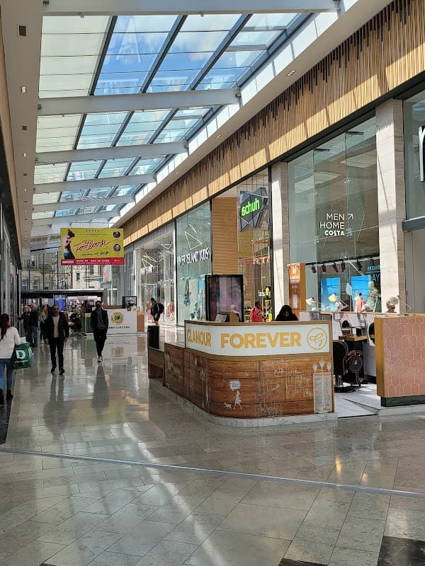 Bright indoor shopping area with shops, a cafรฉ, and a skylight ceiling, bustling with people.