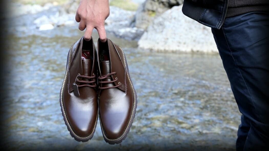 A person holding a pair of brown leather shoes by the laces, standing near a rocky water edge.