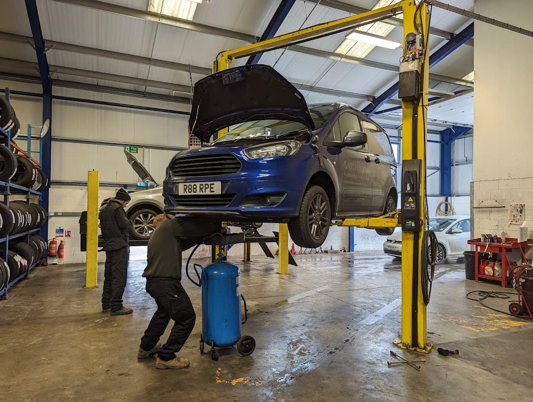 A blue car on a lift in a garage, with a mechanic working underneath and tyres stacked in the background.